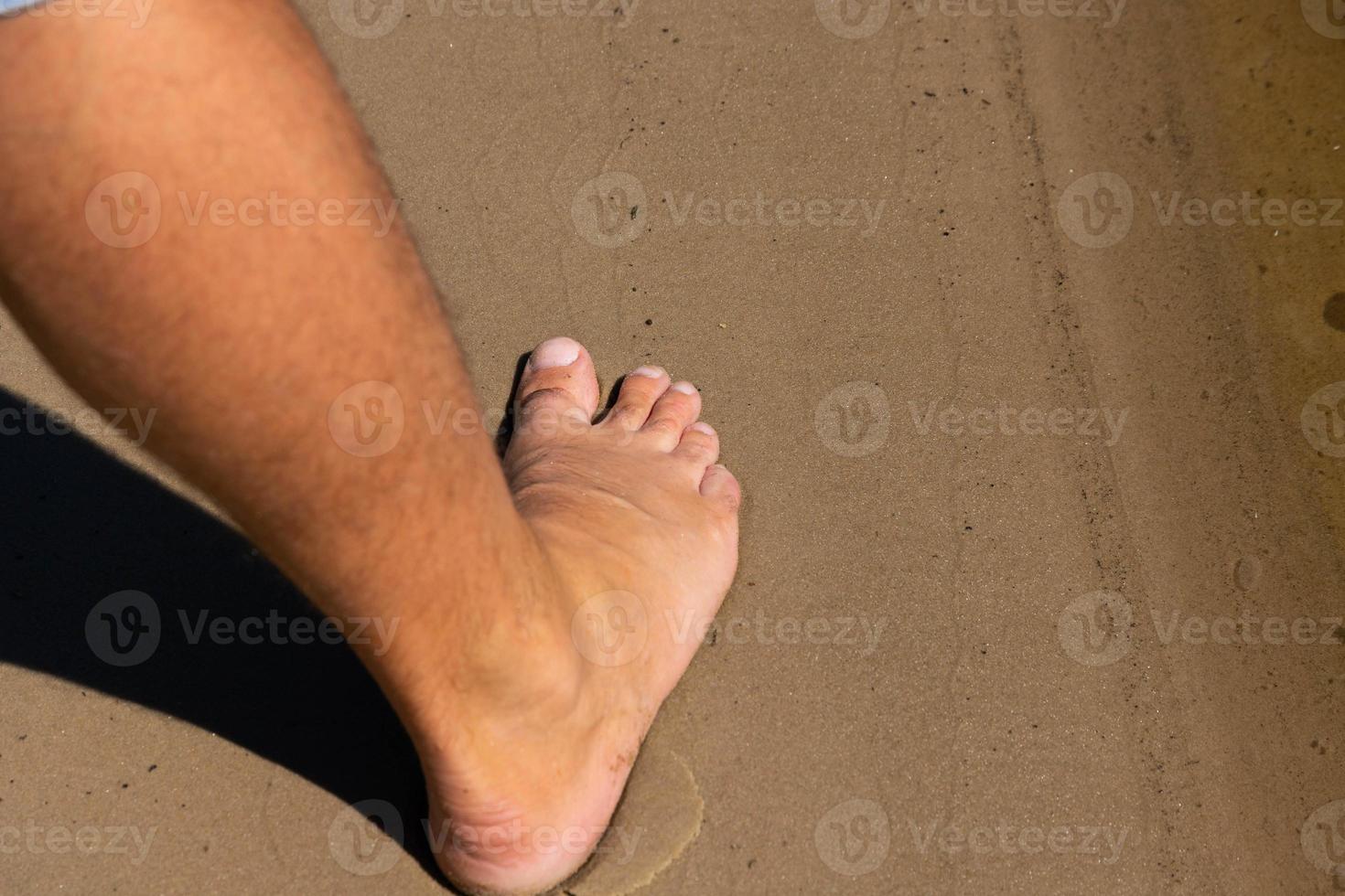 The right leg and foot of a naked man stands on the sand of the coastline. photo