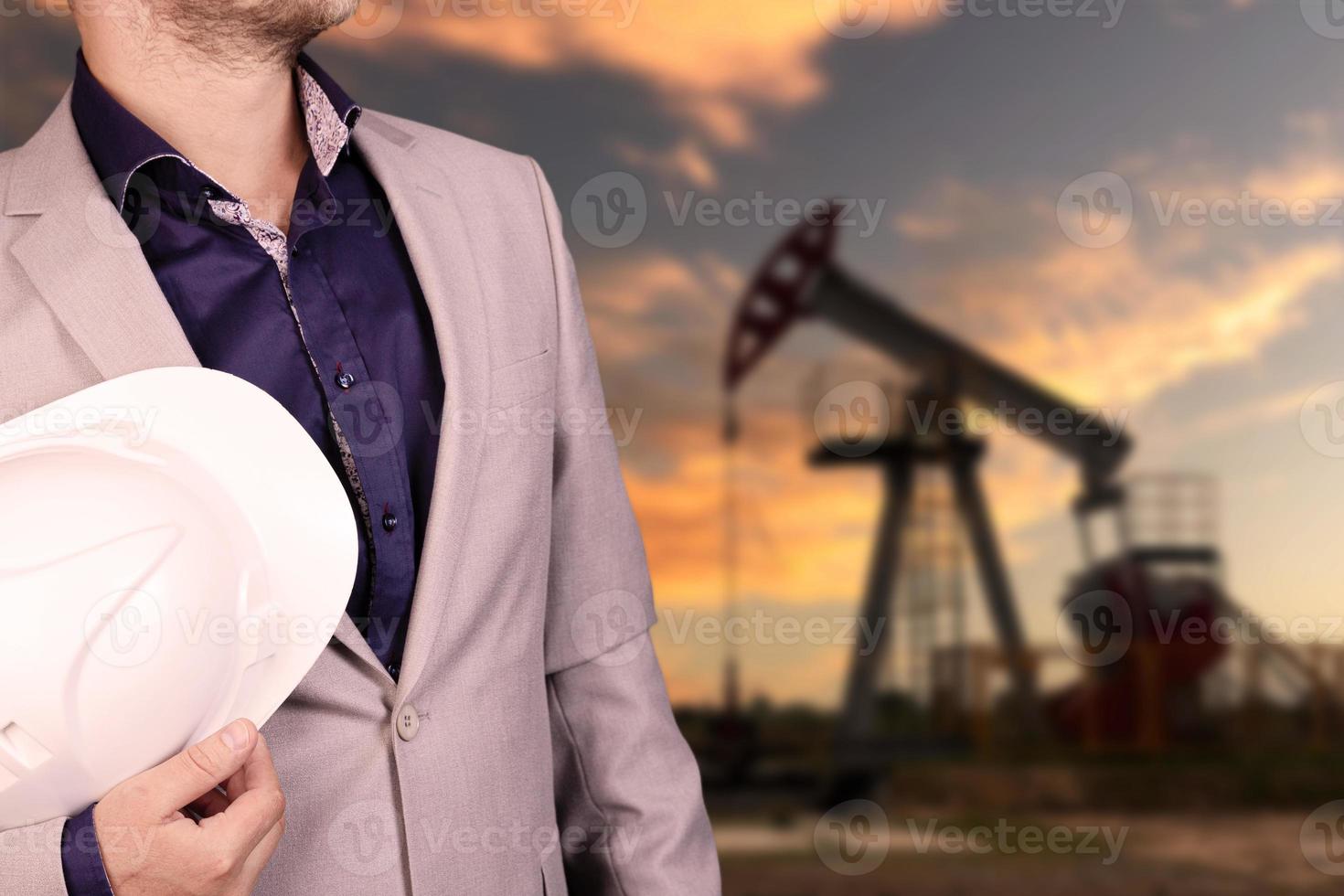 Petroleum engineer working in the oil industry stands with a white helmet in his hands in front of an oil pump. photo