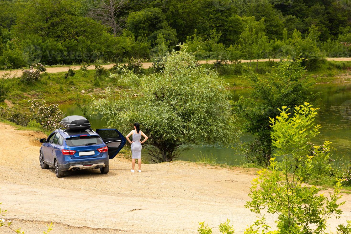 A blue car with a girl with a black roof rack stands against the background of a mountain lake with mountains and clouds. photo