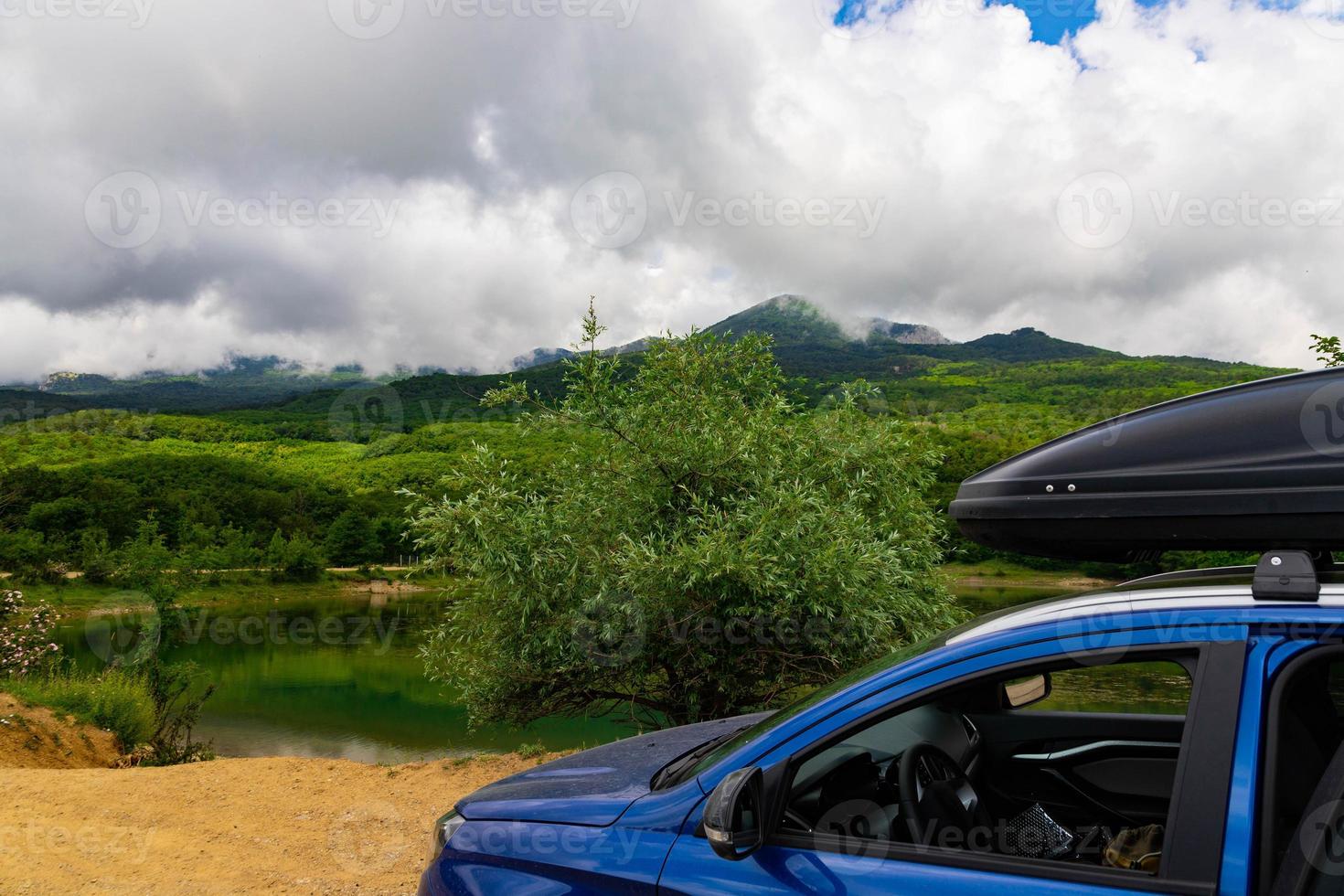 A blue car with a black roof rack stands against the backdrop of a mountain lake with mountains and clouds. photo