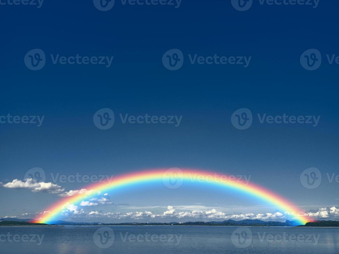 Rainbow on dark blue sky and white cloud on lake and line of tree and tiny mountain photo