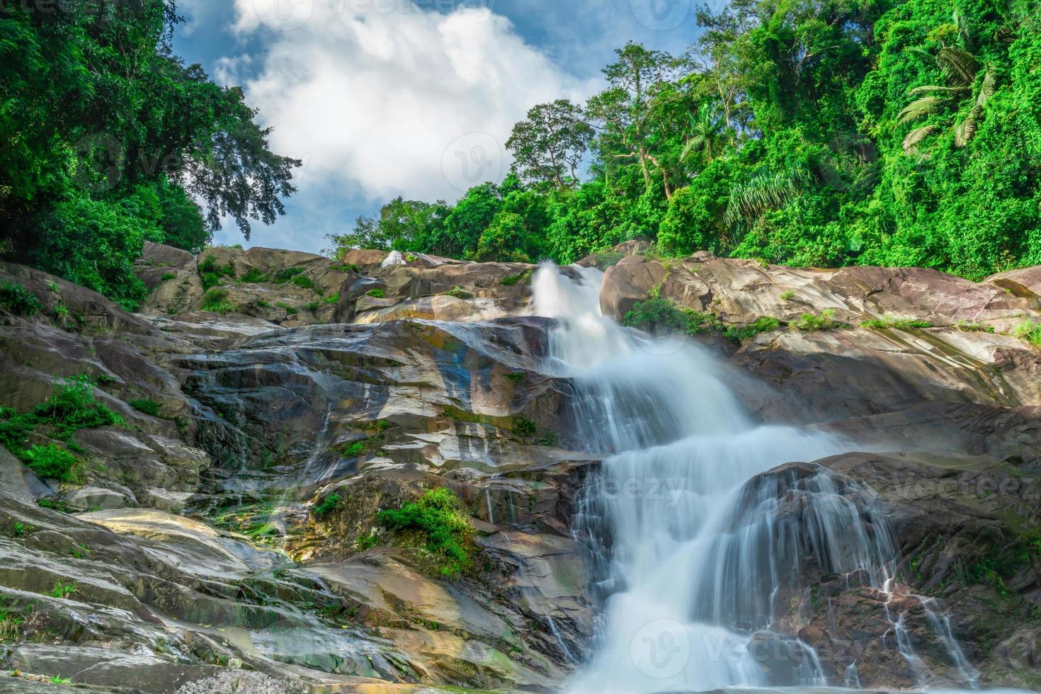 Beautiful waterfall at the mountain with blue sky and white cumulus clouds. Waterfall in tropical green tree forest. Waterfall is flowing in jungle. Nature abstract background. Granite rock mountain. photo