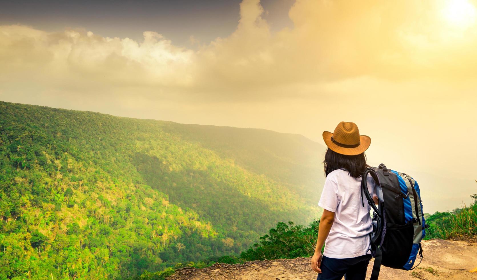 joven viajera con mochila y sombrero en la cima del acantilado de la montaña viendo hermosas vistas de los bosques y el cielo después de la lluvia en sus vacaciones. mujer asiática viaja sola. foto