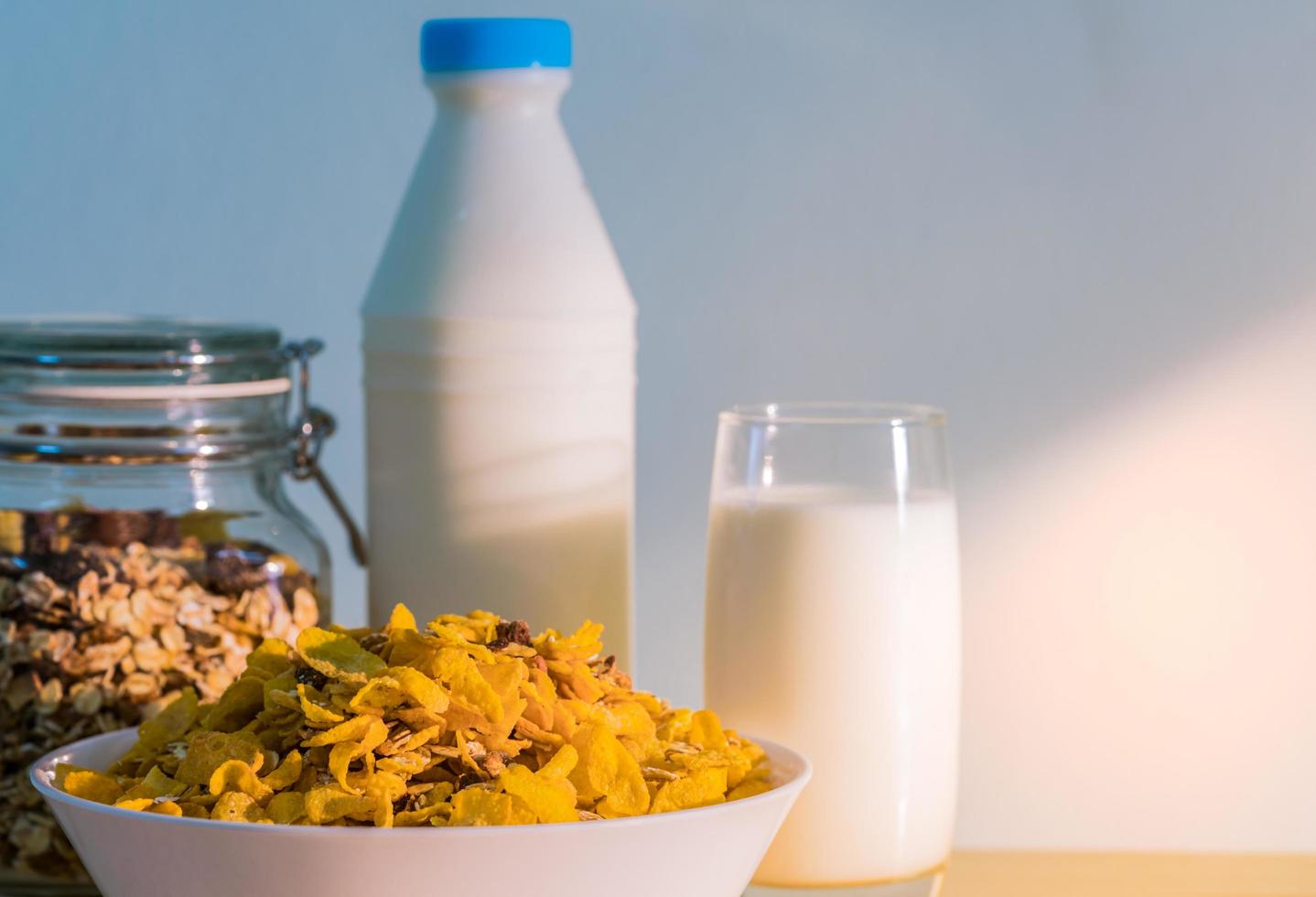 un vaso de leche y una botella de leche con una etiqueta en blanco colocada en una mesa de madera cerca de un tazón de cereal con una cuchara. desayuno de calcio para niños antes de ir a la escuela por la mañana. copos de maíz y concepto de leche foto