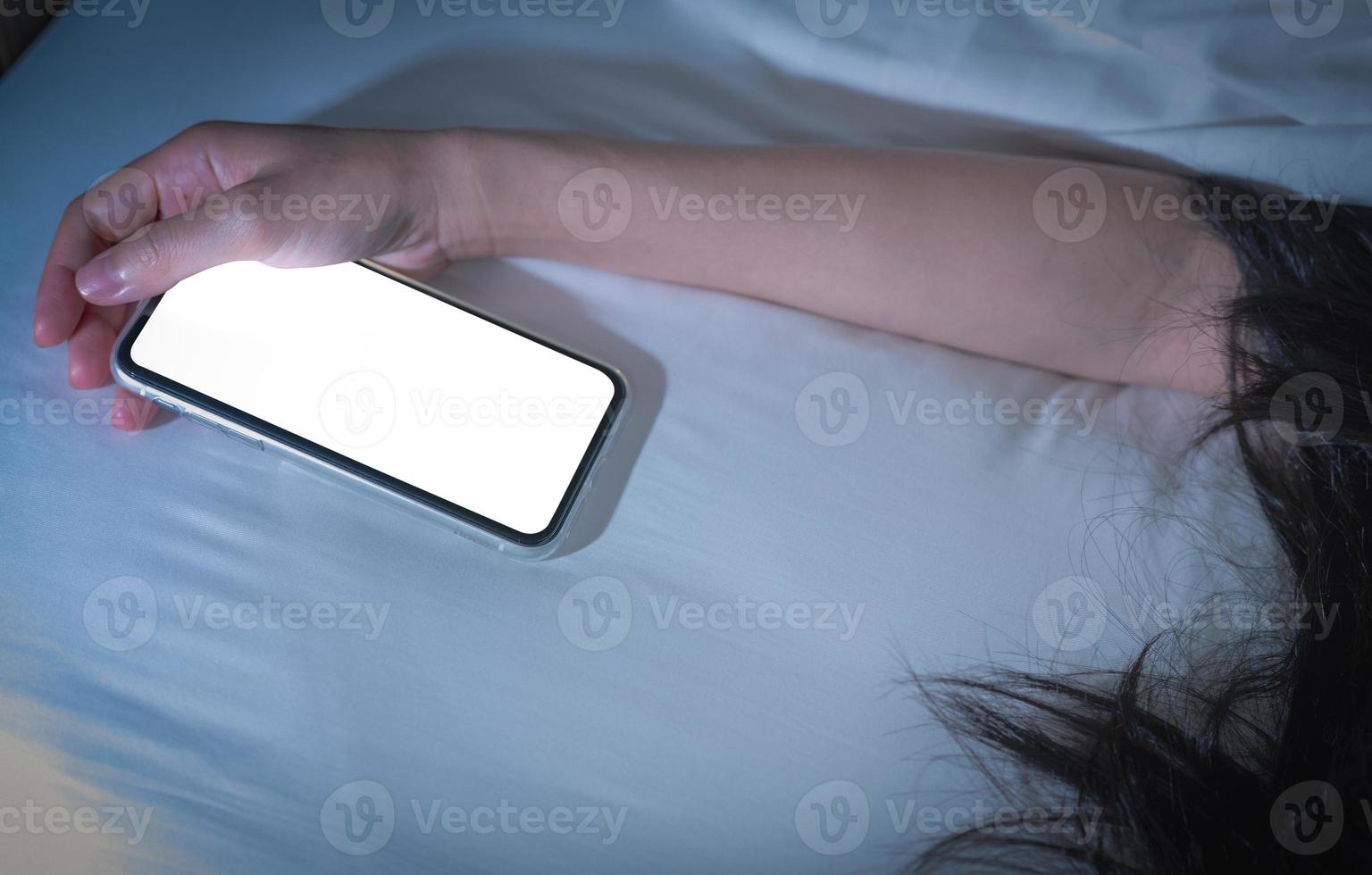 mujer trabajadora durmiendo en la cama en casa y mano sosteniendo un teléfono móvil con pantalla en blanco. mujer usando teléfono inteligente en el dormitorio. mujer joven adicta al teléfono inteligente. enviar mensajes de texto en el teléfono celular con cansancio hasta dormir. foto