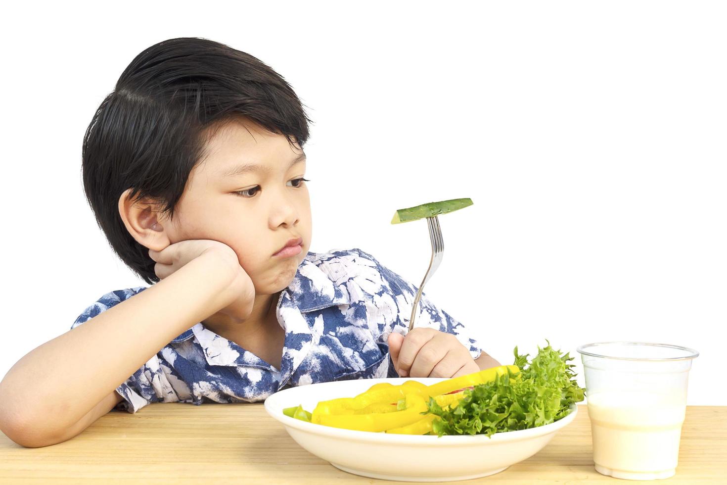 un chico encantador asiático que muestra una expresión aburrida con verduras frescas y coloridas y un vaso de leche aislado sobre fondo blanco foto