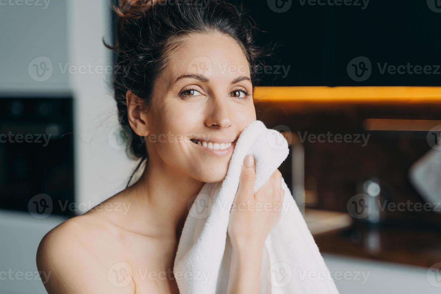 la chica europea se limpia la cara con una toalla después del lavado. joven morena se ducha en casa. foto
