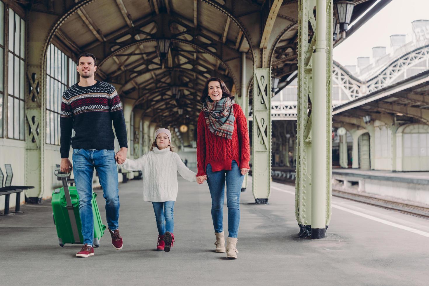 Shot of happy family going to have nice trip during holidays, carry bag, walk on railways station platform, being in good mood. Father, mother and child arrive from journey. Travelling concept photo