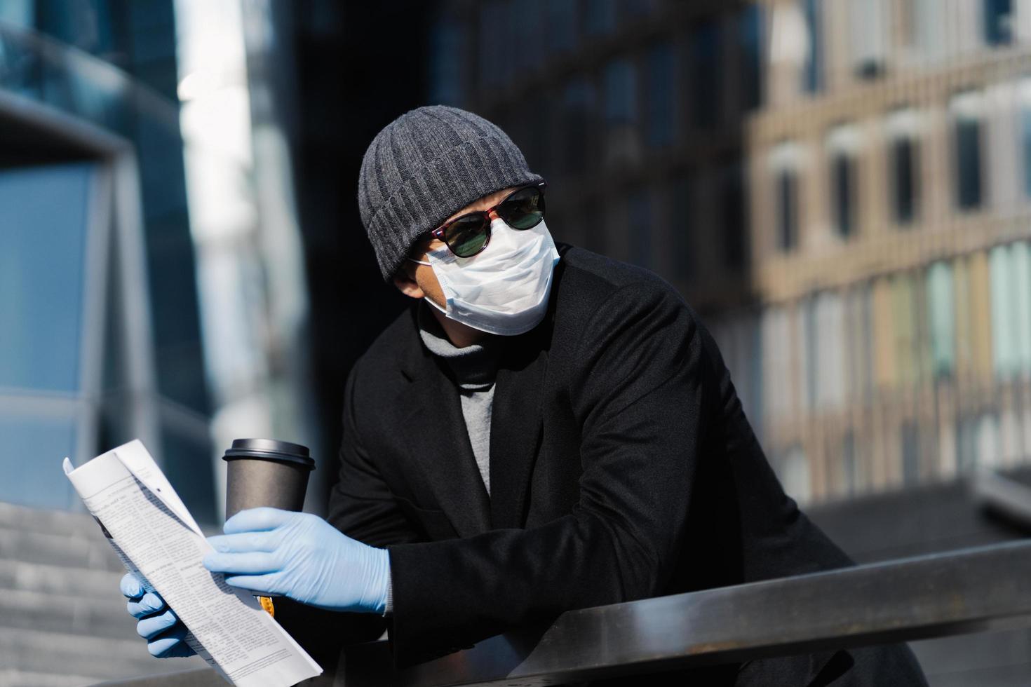 joven en la calle con café para llevar y periódico, usa máscara y guantes médicos protectores, evita la propagación del coronavirus, mira a un lado pensativamente, posa al aire libre. pandemia de covid-19, cuarentena foto