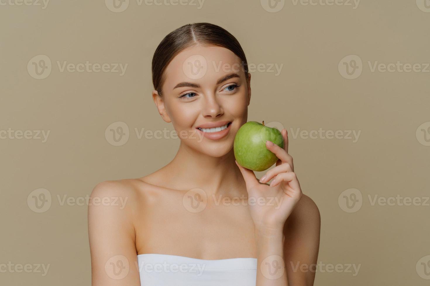 Thoughtful charming European woman holds apple near face smiles gently has white perfect teeth healthy clean skin wrapped in shower towel stands with bare shoulders against brown background. photo