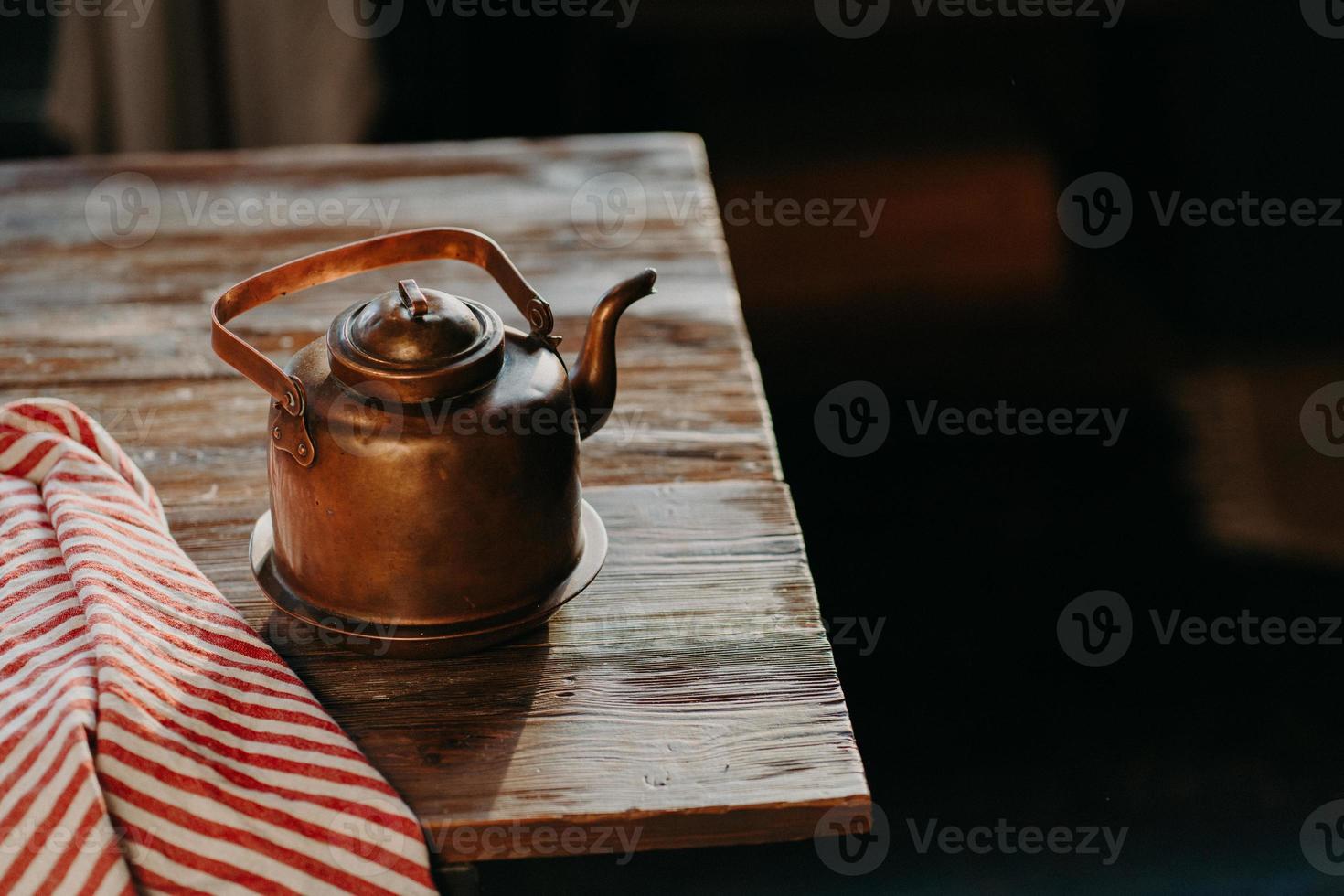 Old copper metal teapot on wooden table in dark room. Red striped towel nearby. Antique kettle for making tea or coffee. Cooking equipment photo