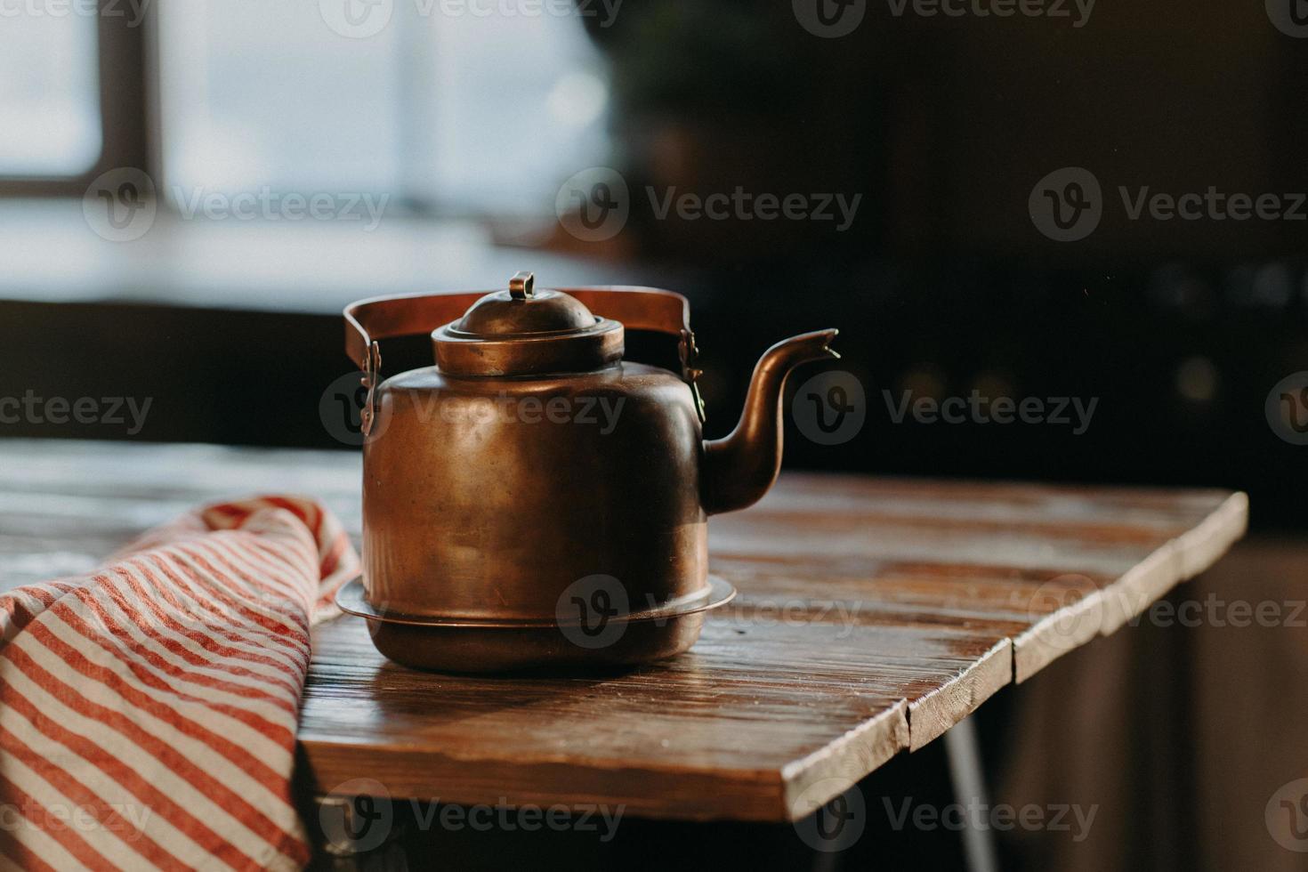 Horizontal shot of aluminium teapot on wooden table against blurred background striped napkin near. Old kettle made of bronze photo