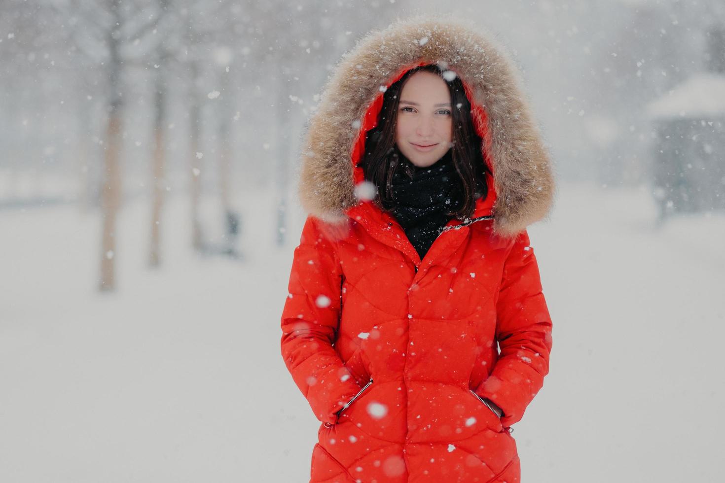 Good looking young woman has appealing look, wears warm red jacket, keeps hands in pockets, has outdoor stroll during winter time and snowfall, poses over blurred snow background. Season concept photo