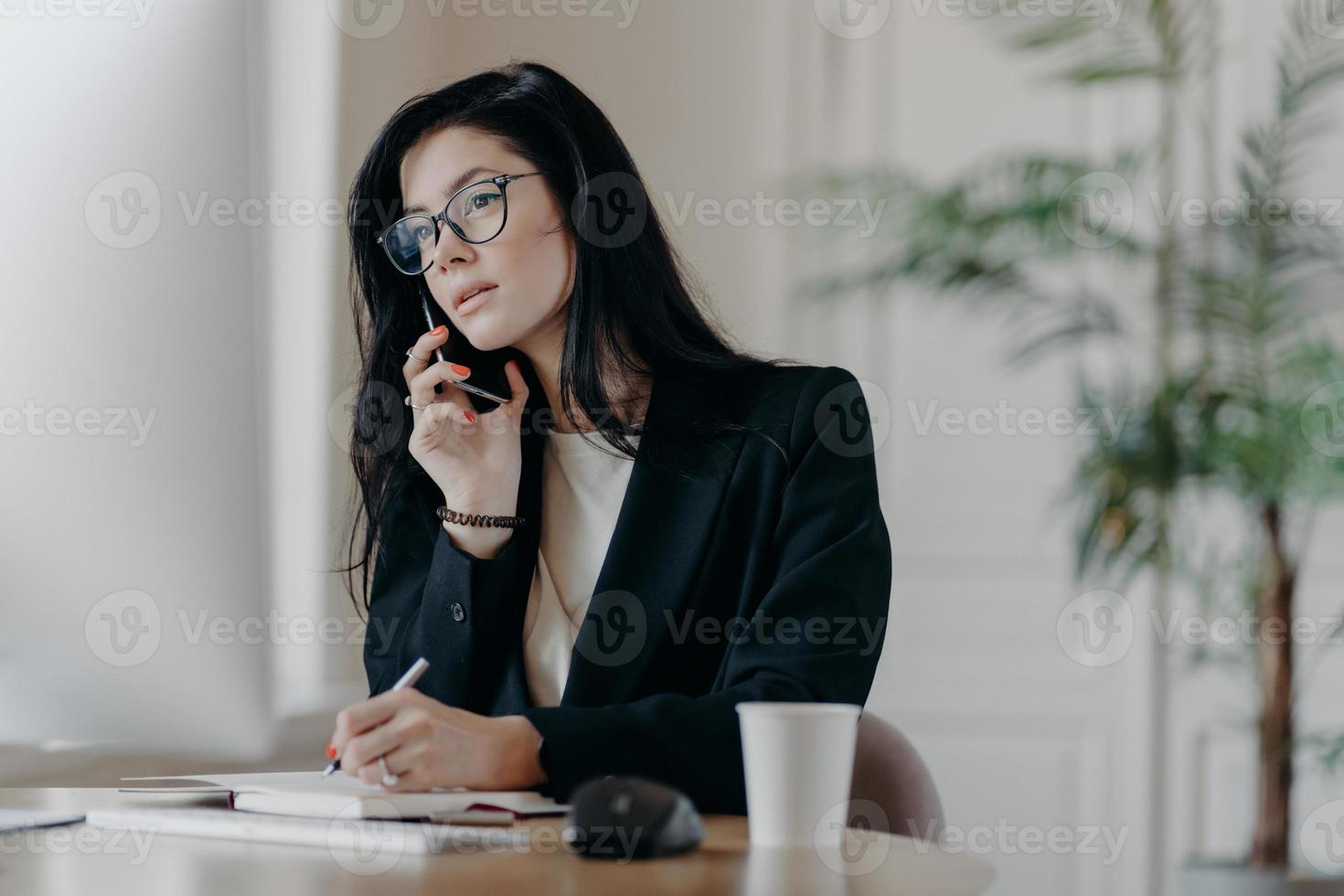 Gorgeous businesswoman makes phone call, concentrated in computer screen, writes down information into notepad, poses at cozy workplace, focused on important task, makes notes in personal organizer photo