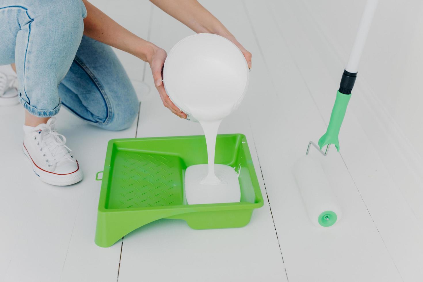 Cropped shot of unrecognizable woman pours white paint in tray, going to paint something with roller, makes house renovation, wears jeans and sneakers. Repair, building, home concept photo