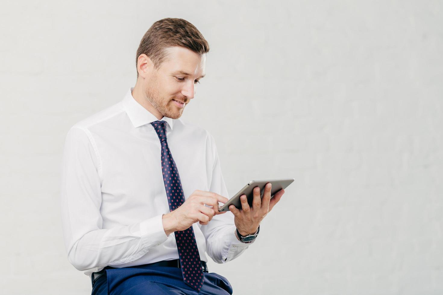 Positive successful male executive manager holds modern touch pad, gets notification about net feeds in social networks, satisfied with high speed internet connection, dressed in formal shirt with tie photo