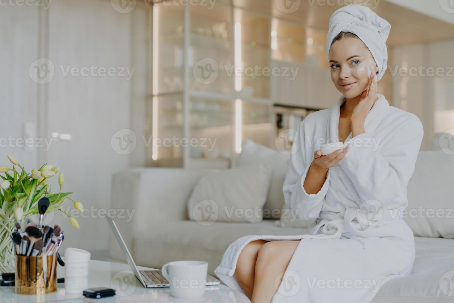 Horizontal shot of pretty Caucasian woman applies facial cream looks thoughtfully aside holds bar of cosmetic product dressed in bathrobe and towel sits on comfortable sofa over home interior photo