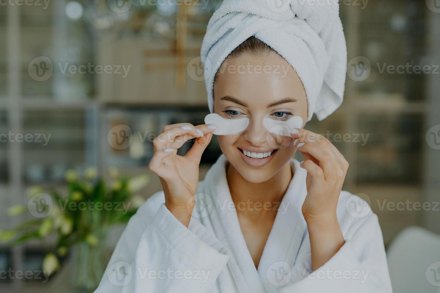 Cheerful young woman with healthy skin takes off beauty patches from under eyes reduces wrinkles or puffiness wears wrapped towel on head and dressing gown smiles toothily has minimal makeup photo