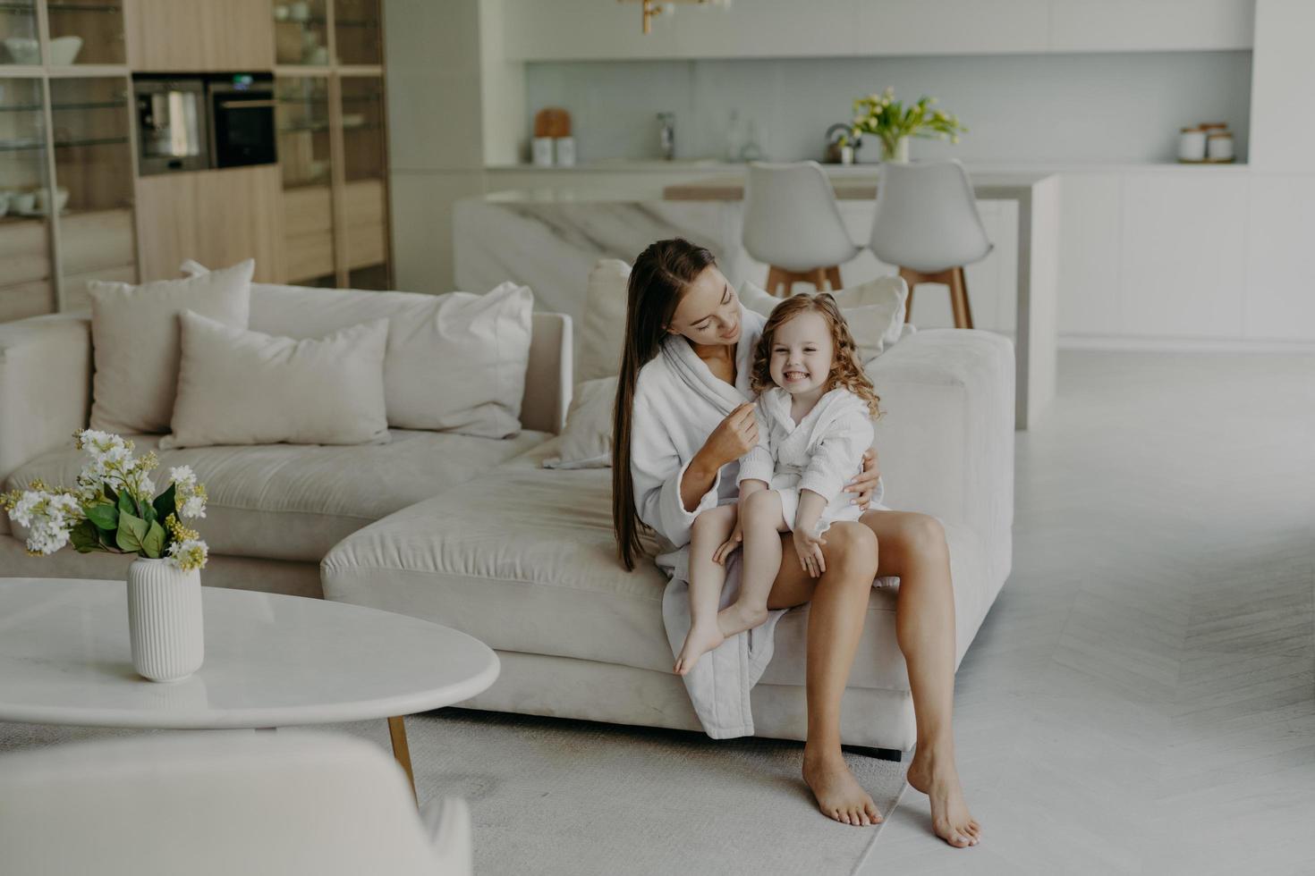 Happy adorable little hands poses on mothers legs dressed in soft white bath towel feels very glad. Mom and daughter wear domestic clothes sit on comfortable sofa in living room. Home interior photo