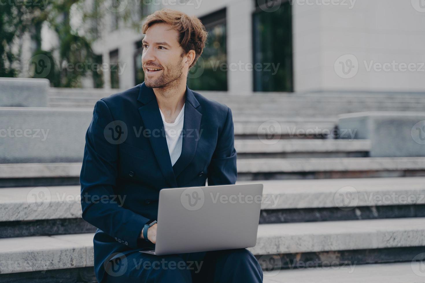 Young smiling office worker sitting outside on stairs in city center holding laptop on his lap photo