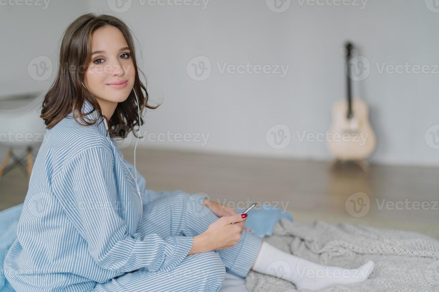 Adorable woman listens lecture on news in internet via cell phone and earphones, has recreation time, connected to wifi at home, poses in bedroom, blurred background with guitar in background photo