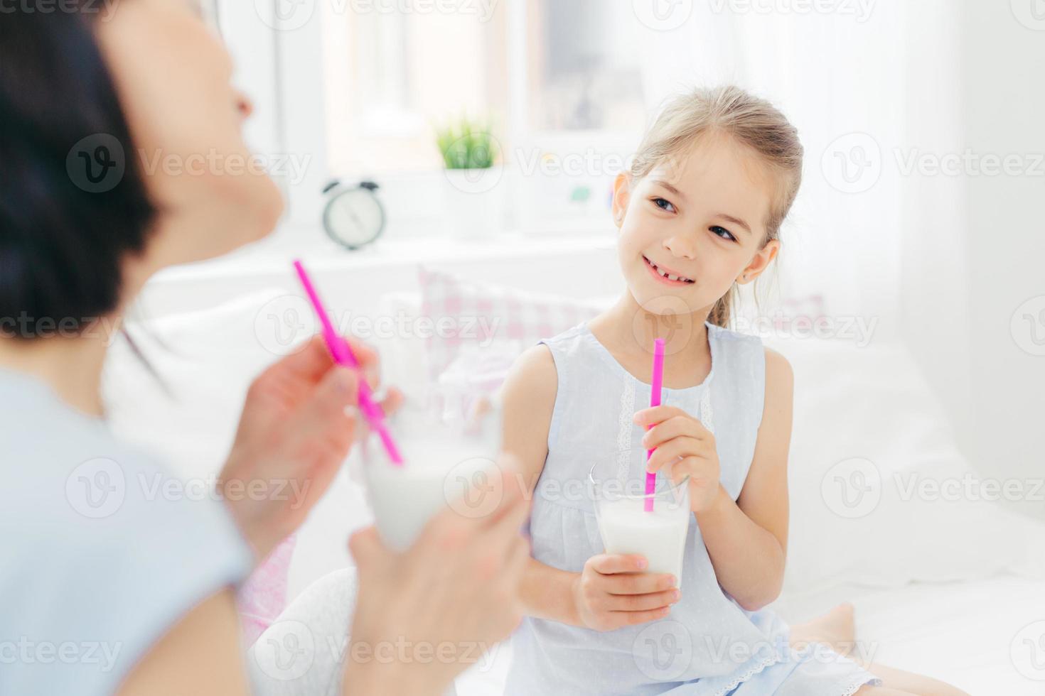 foto recortada de una hermosa niña pequeña bebe un delicioso batido de leche junto con su madre, disfruta de buenos días y un ambiente doméstico tranquilo, tiene una mirada feliz. gente, desayuno y ropa de cama