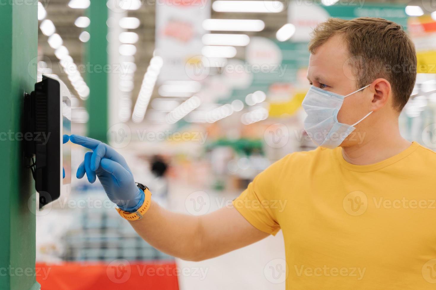 Profile shot of young man types on touch screen of self checkout, wears protective rubber blue gloves, poses against blurred supermarket background. Consumerism, shopping, quarantine concept photo