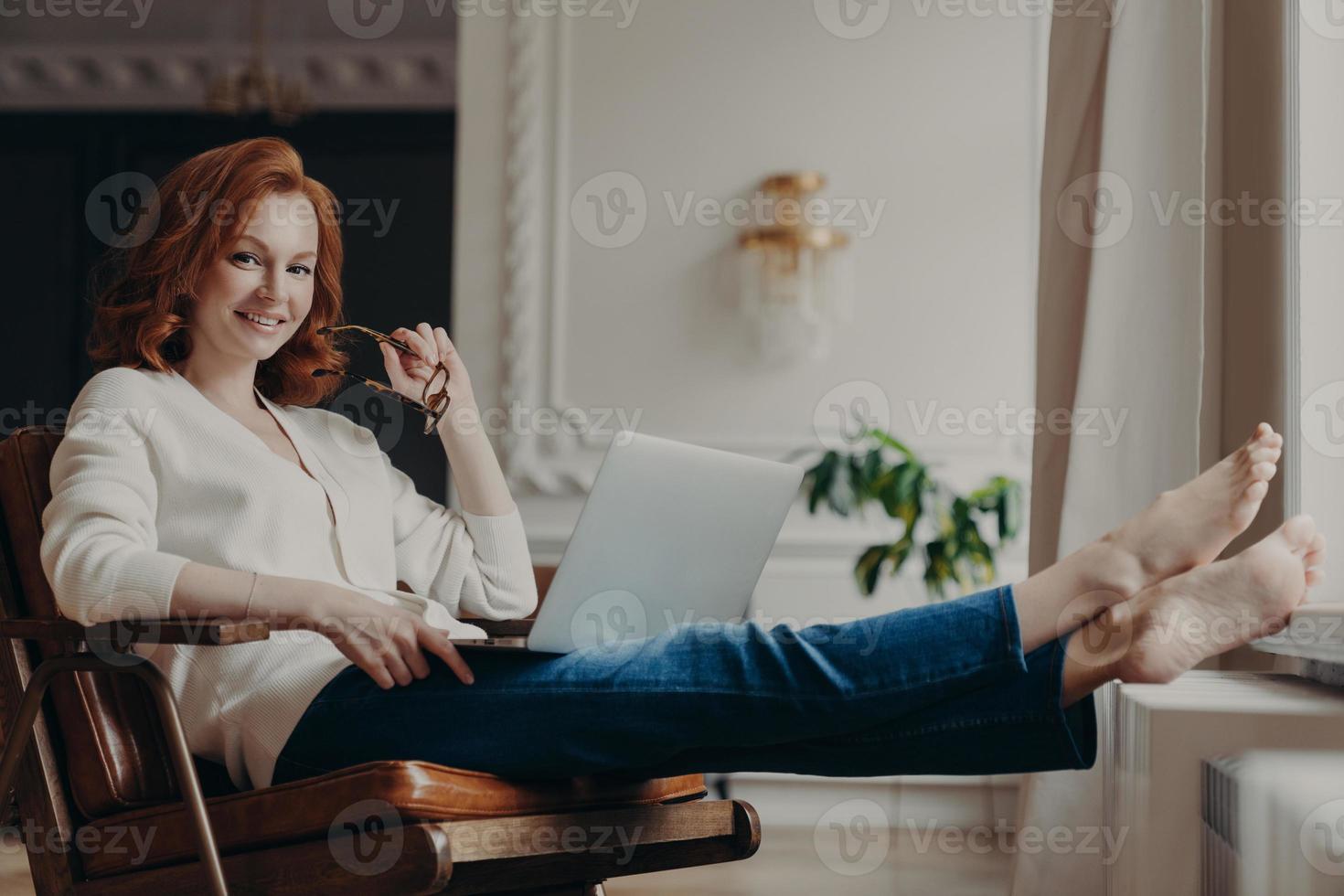 Cheerful successful ginger woman copywriter works from home, keeps laptop on knees, stretches legs, thinks on idea for creating advertising content on own website, browses information in internet photo