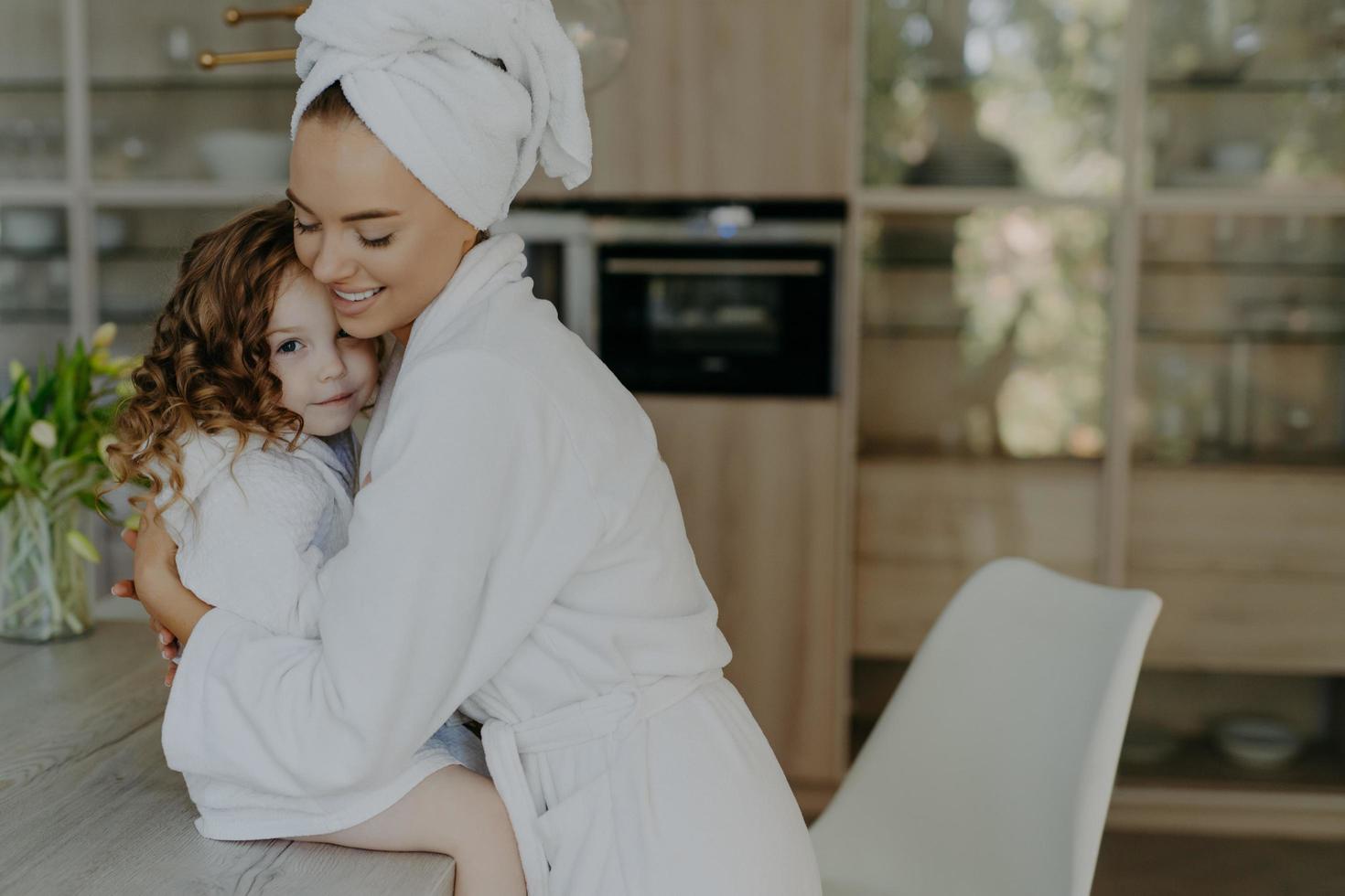 Happy affectionate young mother in bath towel and dressing gown embraces with love who sits at table feel refreshed after taking shower spend free time at home. Family beauty wellness concept photo