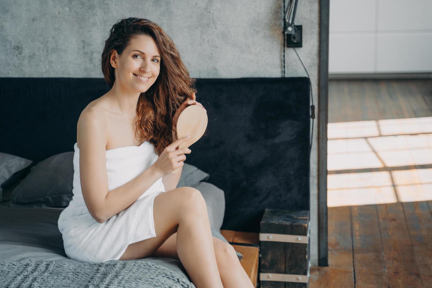 hermosa mujer europea envuelta en toalla peina su largo cabello sedoso con cepillo y sonriendo. foto