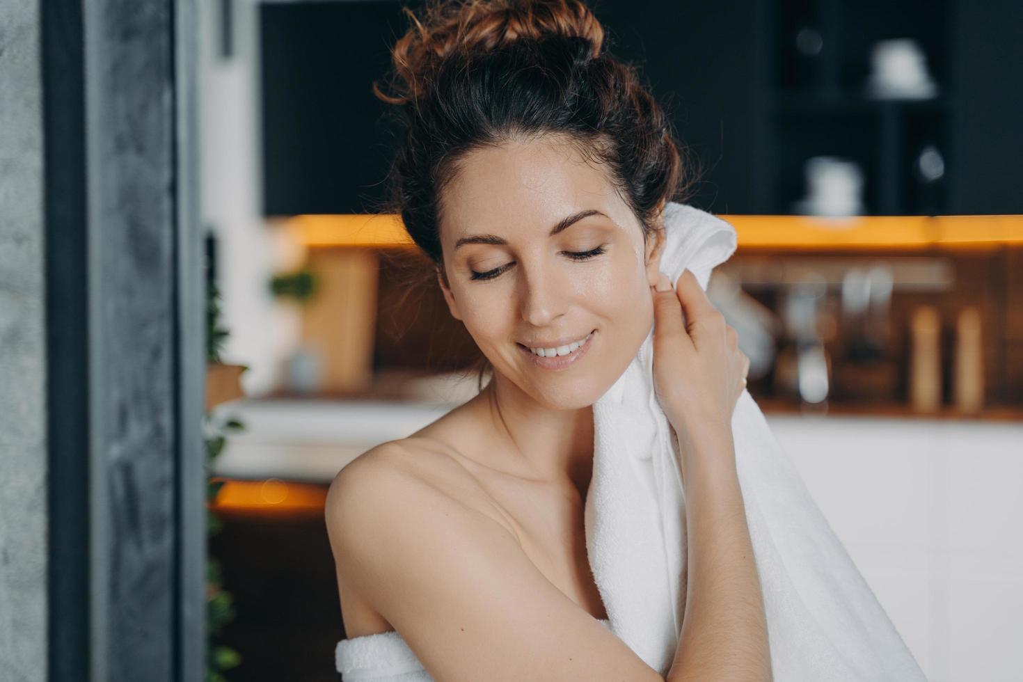 Young lady relaxing in bathroom, enjoying the bath and beauty routine. Girl wiping face with towel. photo