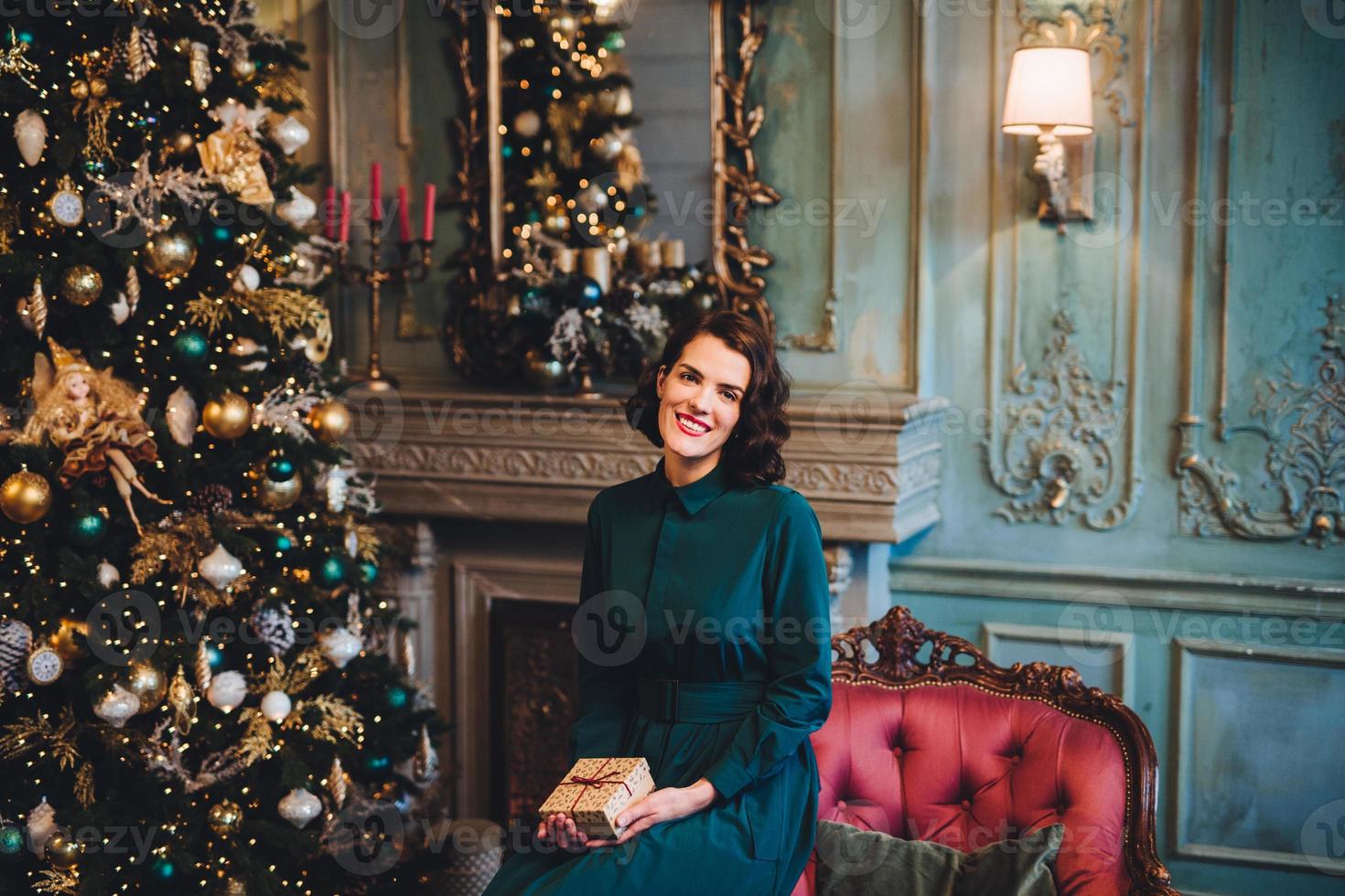 People, celebration, holidays concept. Smiling beautiful woman wears nice dress, sits near Christmas tree in room, holds present, glad to celebrate her favourite holiday. Merry Christmas and New Year photo