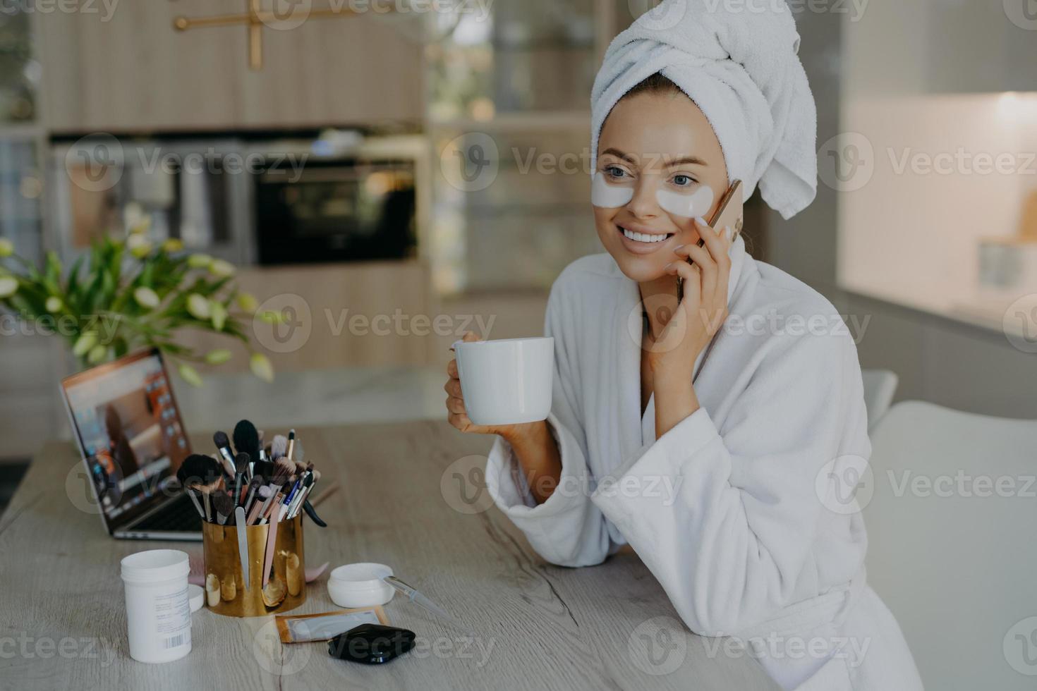 Beauty treatment personal care and hygiene concept. Lovely young woman applies cosmetic patches under eyes wears bathrobe drinks coffee talks to friend via smartphone sits at table over home interior photo