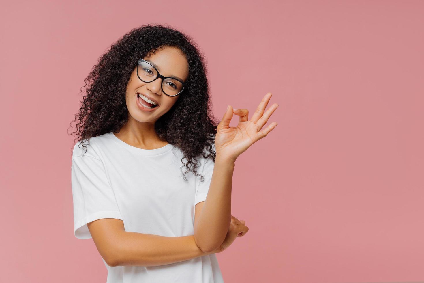 una mujer encantadora y optimista de piel oscura hace un buen gesto, inclina la cabeza, demuestra aprobación, está de acuerdo con algo, tiene una expresión feliz, usa anteojos y una camiseta blanca informal, aislada en una pared rosa foto