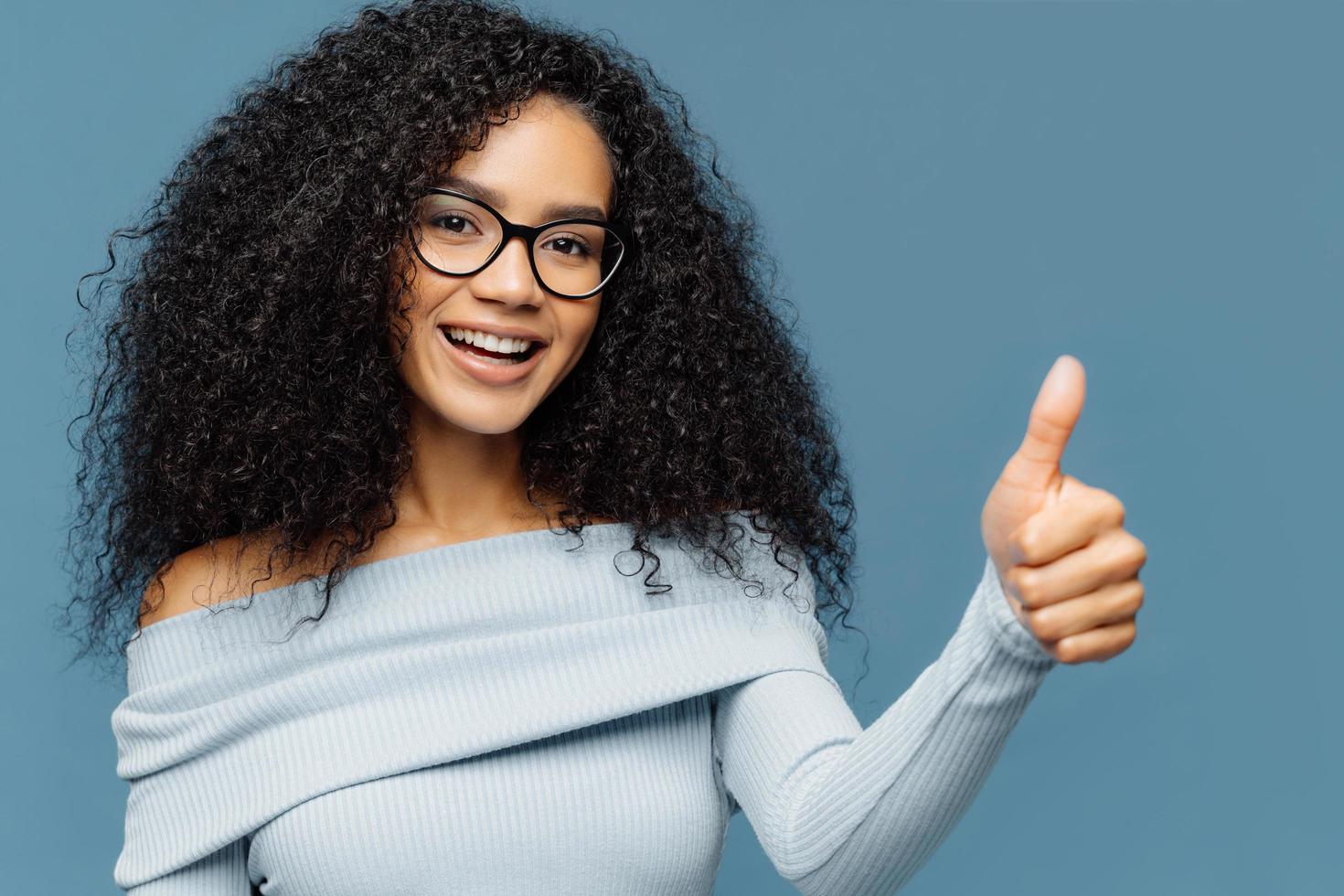 Horizontal shot of lovely dark skinned woman gives thumb up, wears fashionable sweater, eyewear, demonstrates approval, isolated on blue background. Nice choice, good job, well done concept. photo