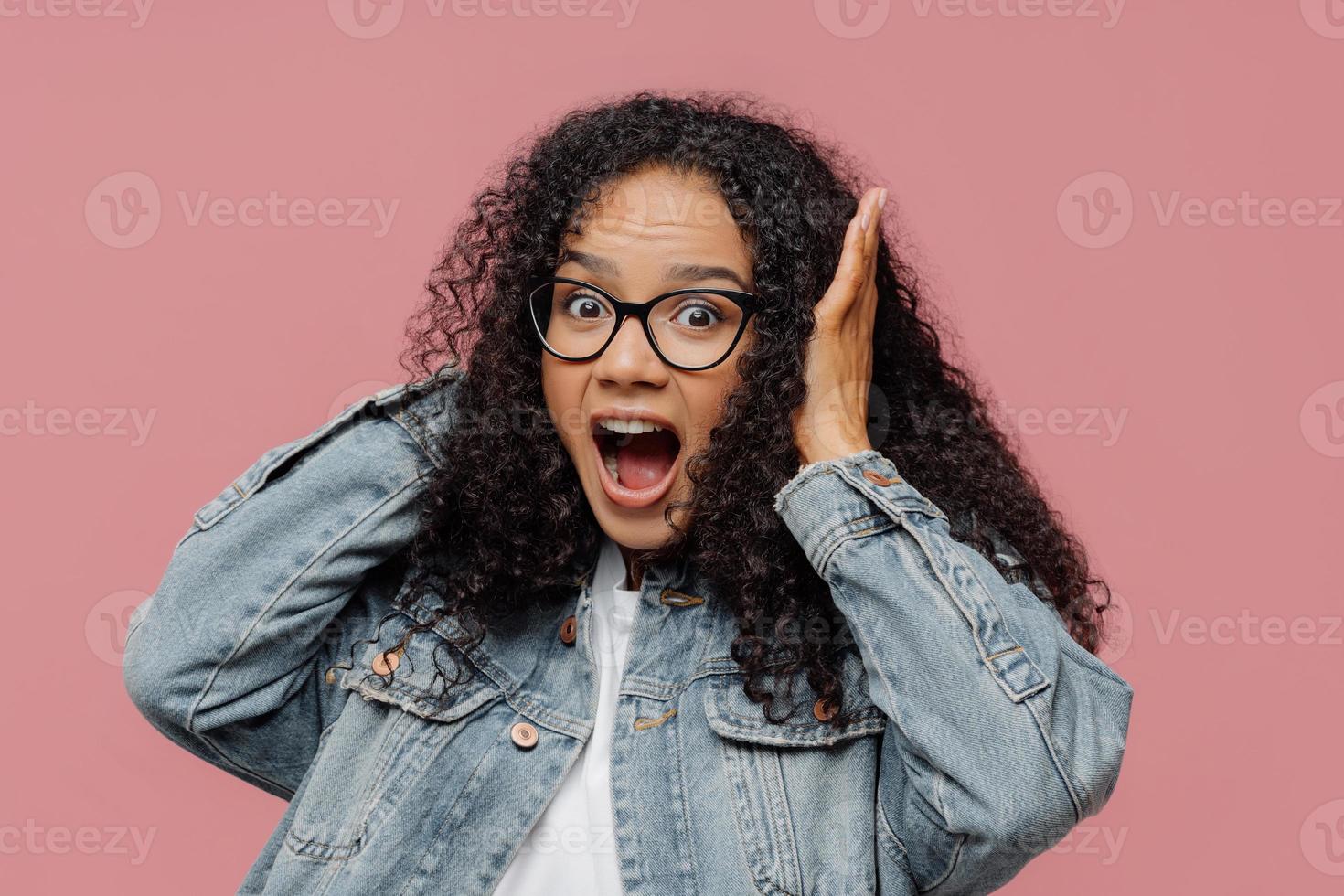 Photo of surprised Afro American woman covers ears, yells loudly, ignores loud sound, keeps mouth widely opened, wears spectacles and denim jacket, stands against rosy wall. Omg, how noisy there