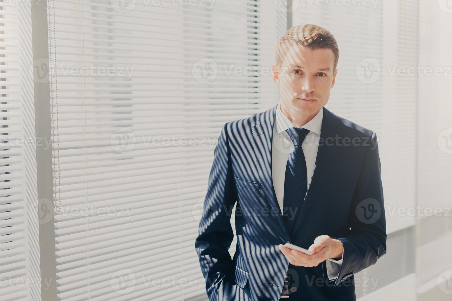 Candid shot of prosperous businessman checks notification on cellular, plans daily schedule, wears formal suit, stands near window, looks directly into camera, has work break. Office routine photo