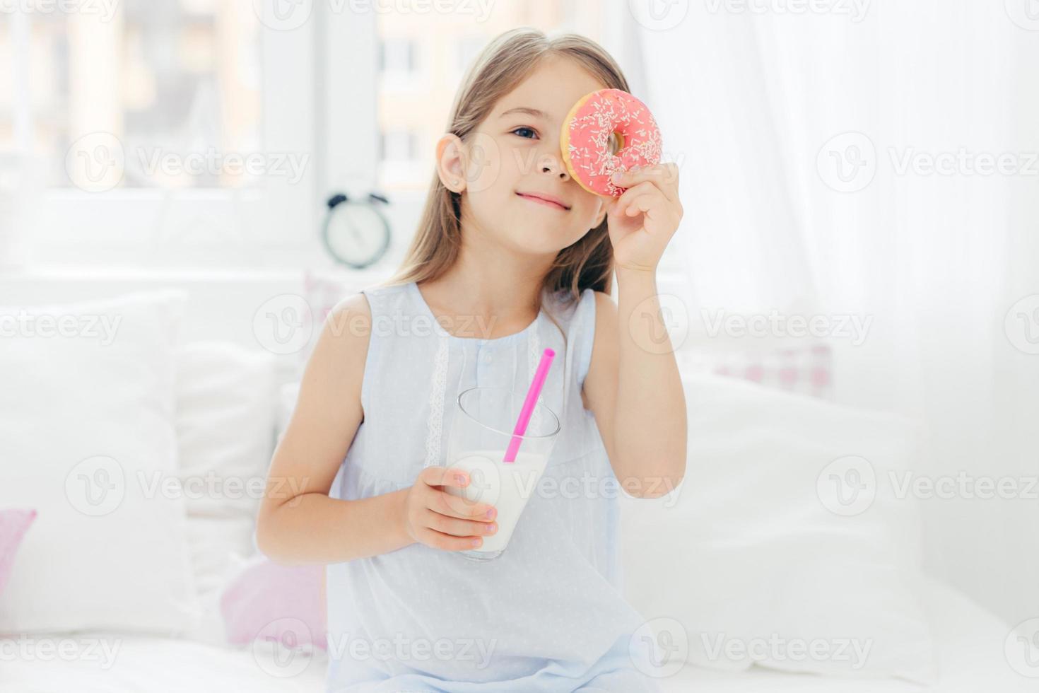 Cheerful little female child poses in bedroom wih delicious doughnut and milk shake, sits on comfortable bed, has fun in morning, going to have tasty breakfast. Children and bed time concept photo