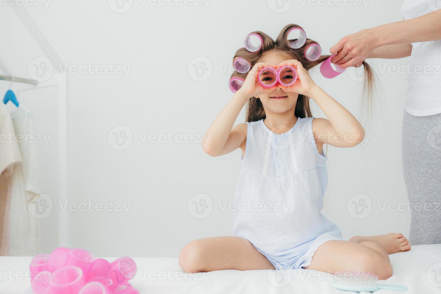 Horizontal shot of beautiful small kid has curlers on head, plays with them, going to have wonderful hairstyle on carnival, isolated over white background. Funny daughter plays together with mum photo