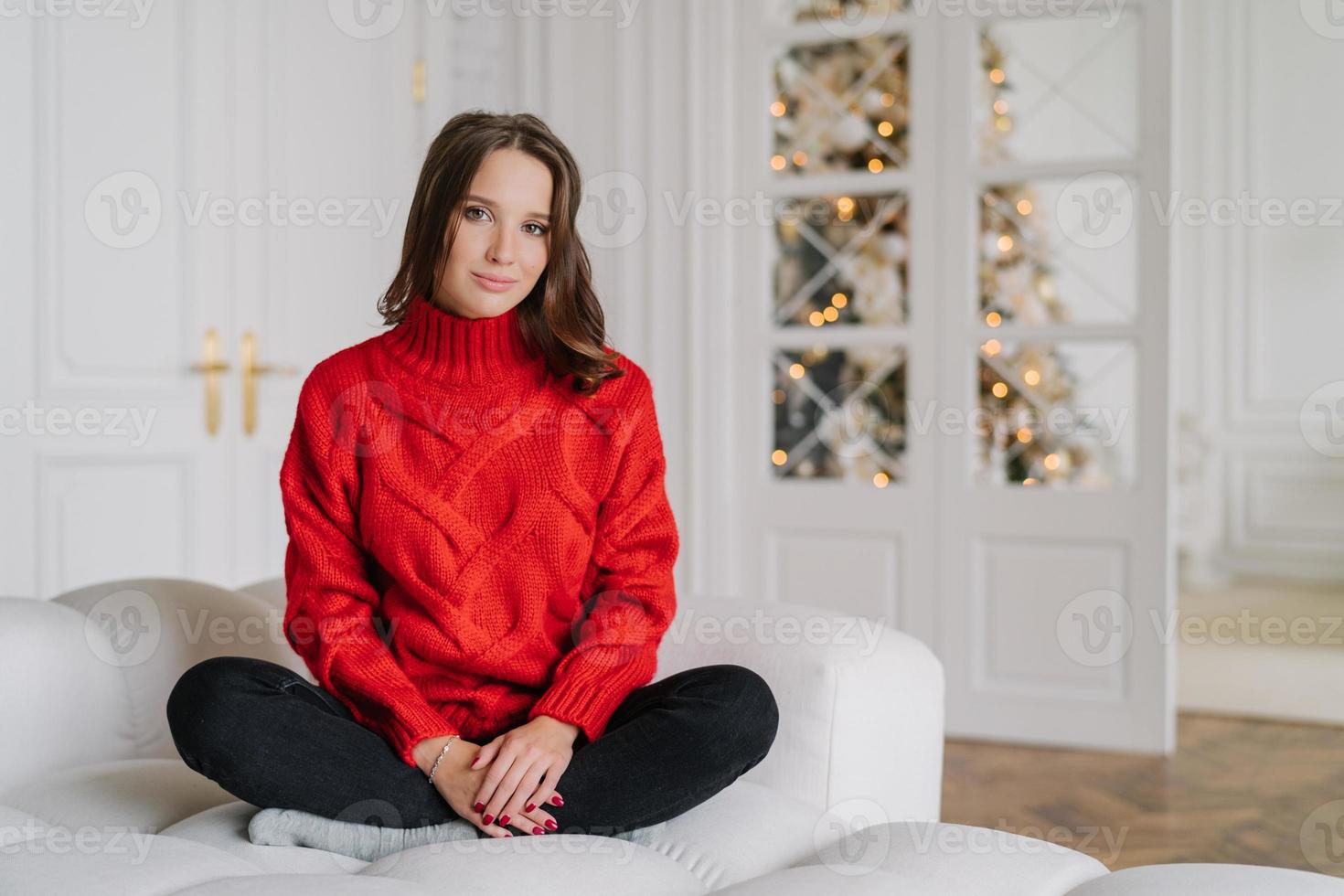Horizontal shot of relaxed housewife dressed in warm red knitted sweater, sits crossed legs in lotus pose on couch, in spaciouss room with New Year tree in background, light colours. Rest concept photo