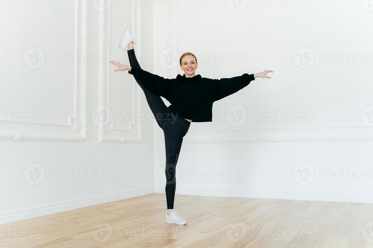 Satisfied female ballerina balances on one leg, stretches in studio, practices yoga, demonstrates good flexibility, dressed in black clothes, has happy expression, performes exercise indoor. photo