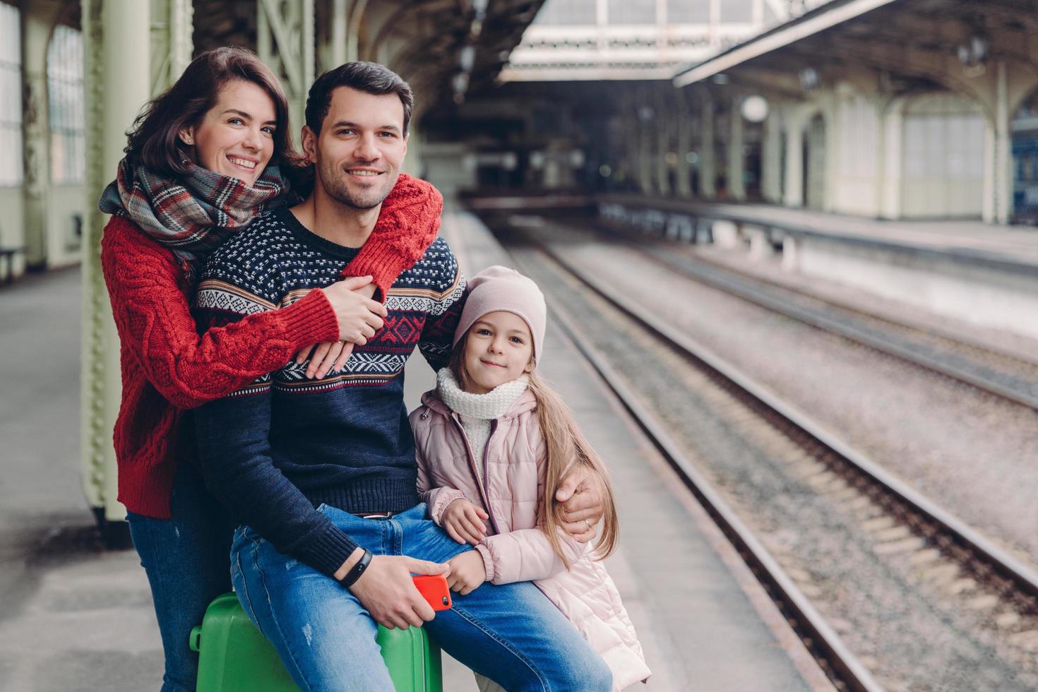 foto de una familia amistosa que tiene una buena relación, tiene un viaje durante las vacaciones, posa en la plataforma de la estación de tren. una mujer encantadora abraza a su marido, su pequeña hija se para cerca de los padres cerca del equipaje