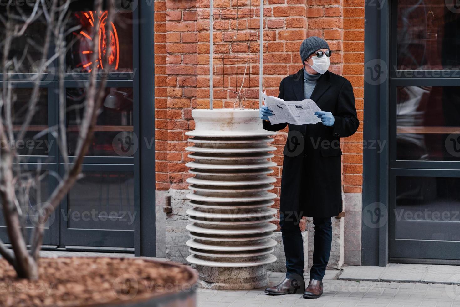Horizontal shot of young man looks aside, wears medical rubber gloves and face mask, looks aside, holds opened newspaper, reads news, thinks about virus protection. Coronavirus outbreak concept photo