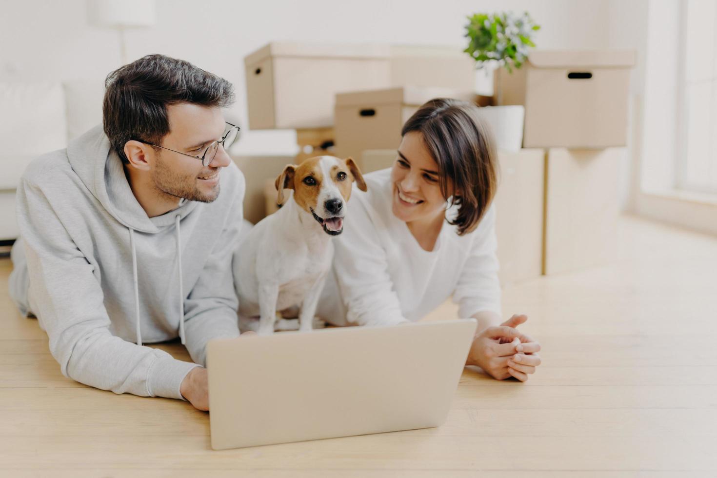 Lovely just married couple lie on floor, use laptop computer, creats design project for new house, their favourite dog poses between them, pile of cardboard boxes in background. Moving to new home photo