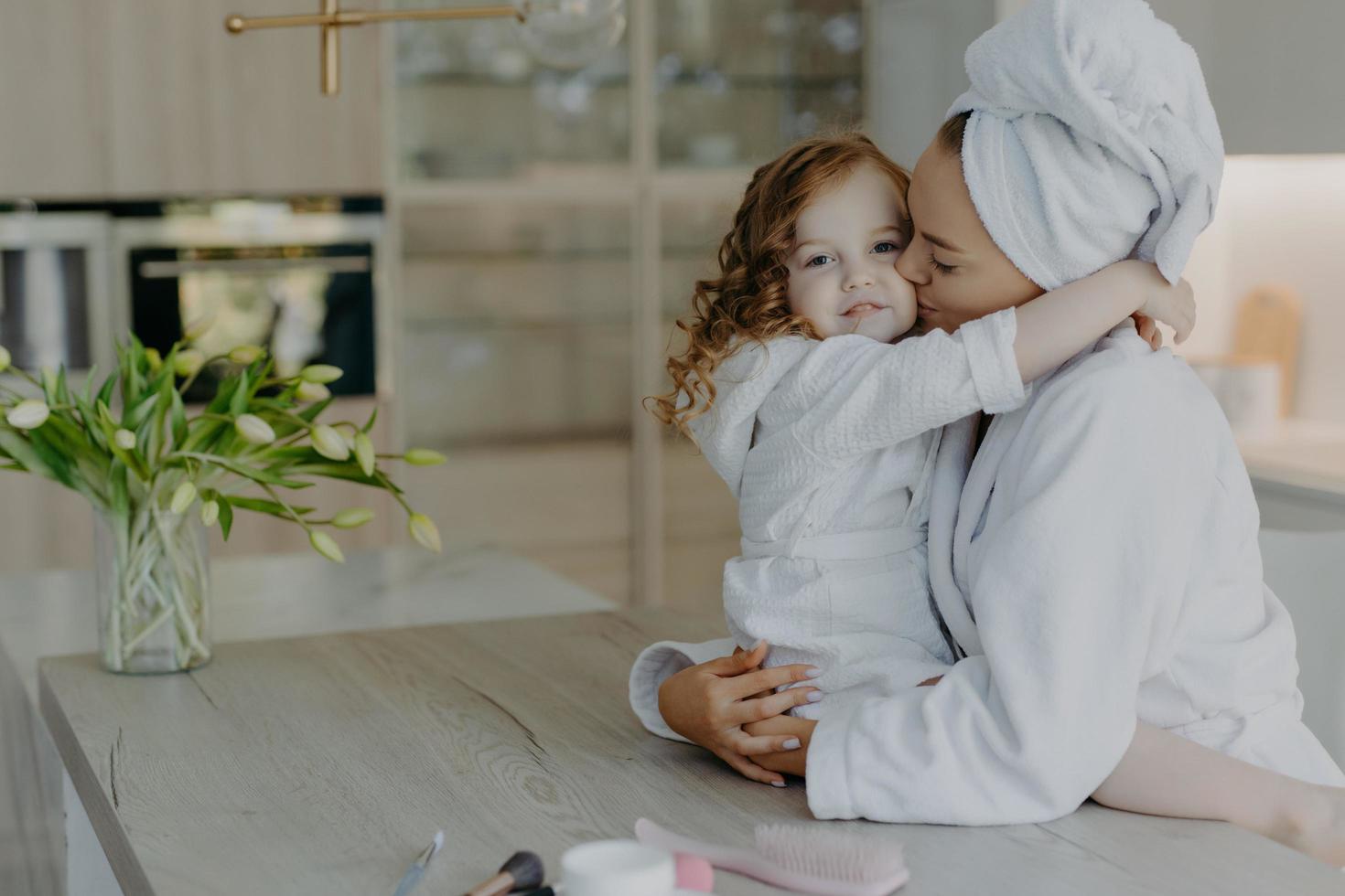 Affectionate mother wearing dressing gown wrapped bath towel on head embraces and kisses with love her small daughter pose together at home against cozy interior feel refreshed after taking shower photo