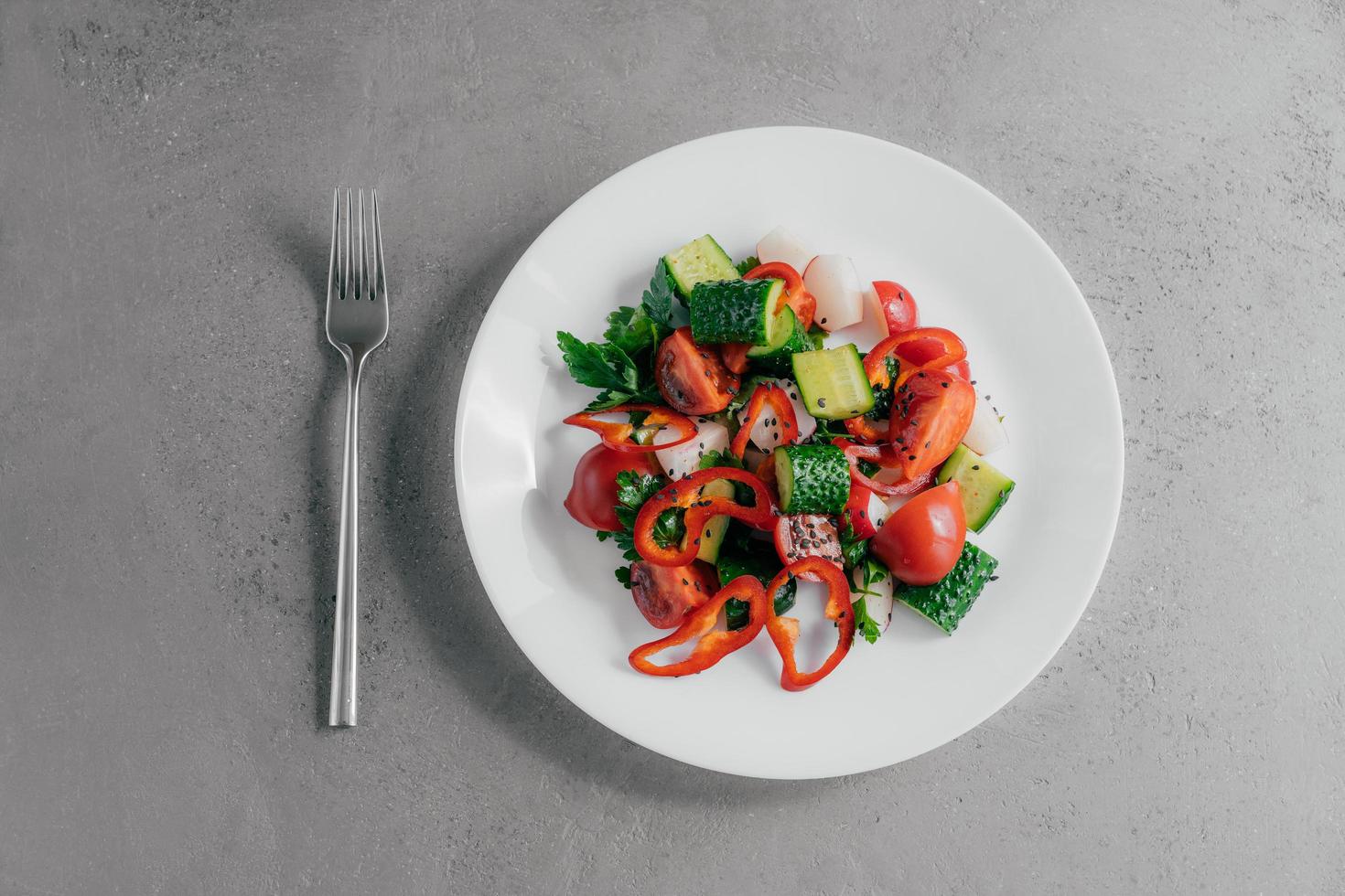 Top view of fresh vegetable salad prepared of red pepper, radish, tomatoes, cucumbers and parsley in white bowl, fork near. Vegetarian dish concept. Healthy nutritious spring salad photo