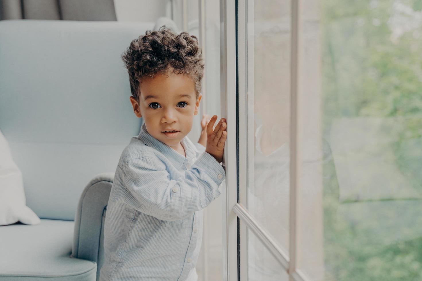 Portrait of cute mixed race curly haired baby boy waiting for parents near window photo