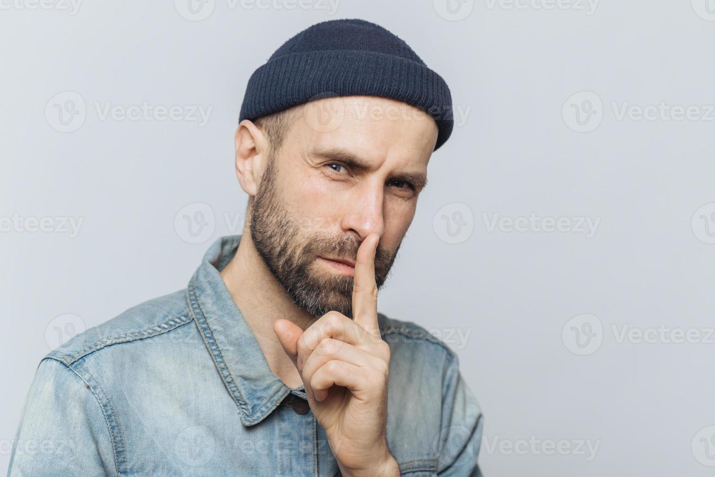 Portrait of serious bearded male with attractive look, keeps fore finger on lips, looks with secret expression, asks to keep silence and not tell his secret to anybody, isolated over grey background photo