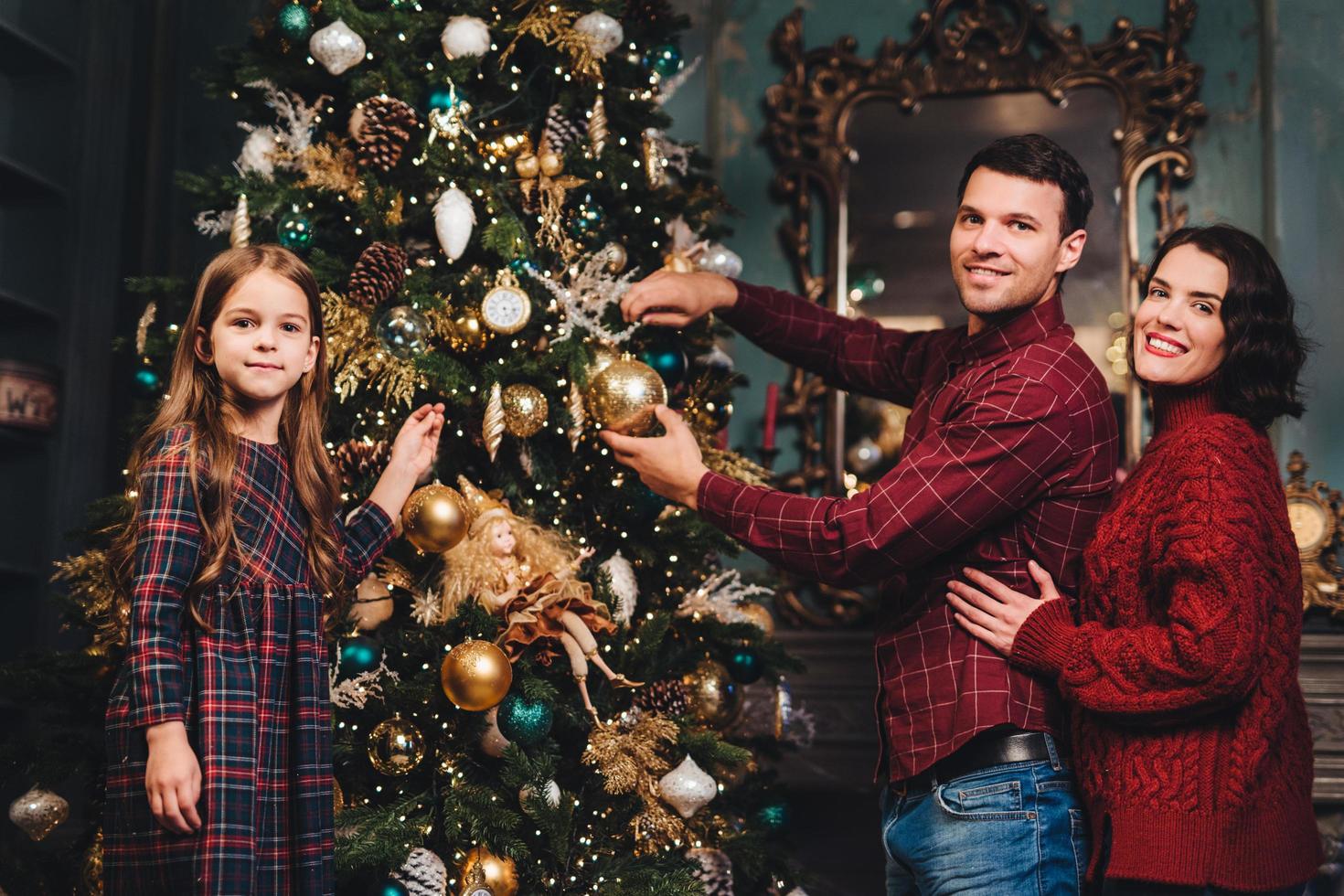 Friendly family of father, mother and daughter decorates New Year tree, being glad to have coming winter holidays, enjoy domestic atmosphere. Family of three members being busy with decorations photo
