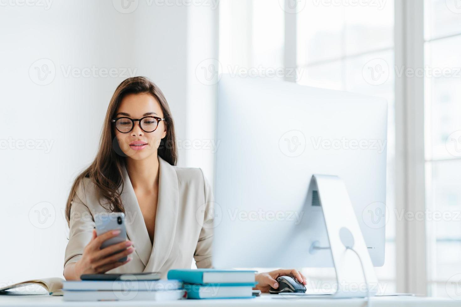 Elegant woman office worker uses mobile phone and computer at one time, sends messages and chats with clients online, monitors news from networks, sits at desktop with pile of notepads and books photo
